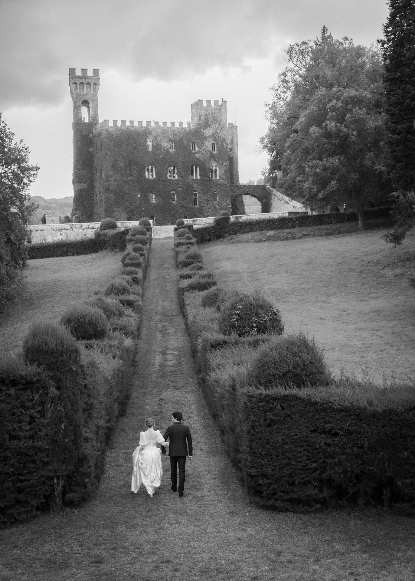 Bride and groom walking hand in hand through the gardens of Castello di Celsa, Tuscany, captured in an elegant black and white wedding photograph.