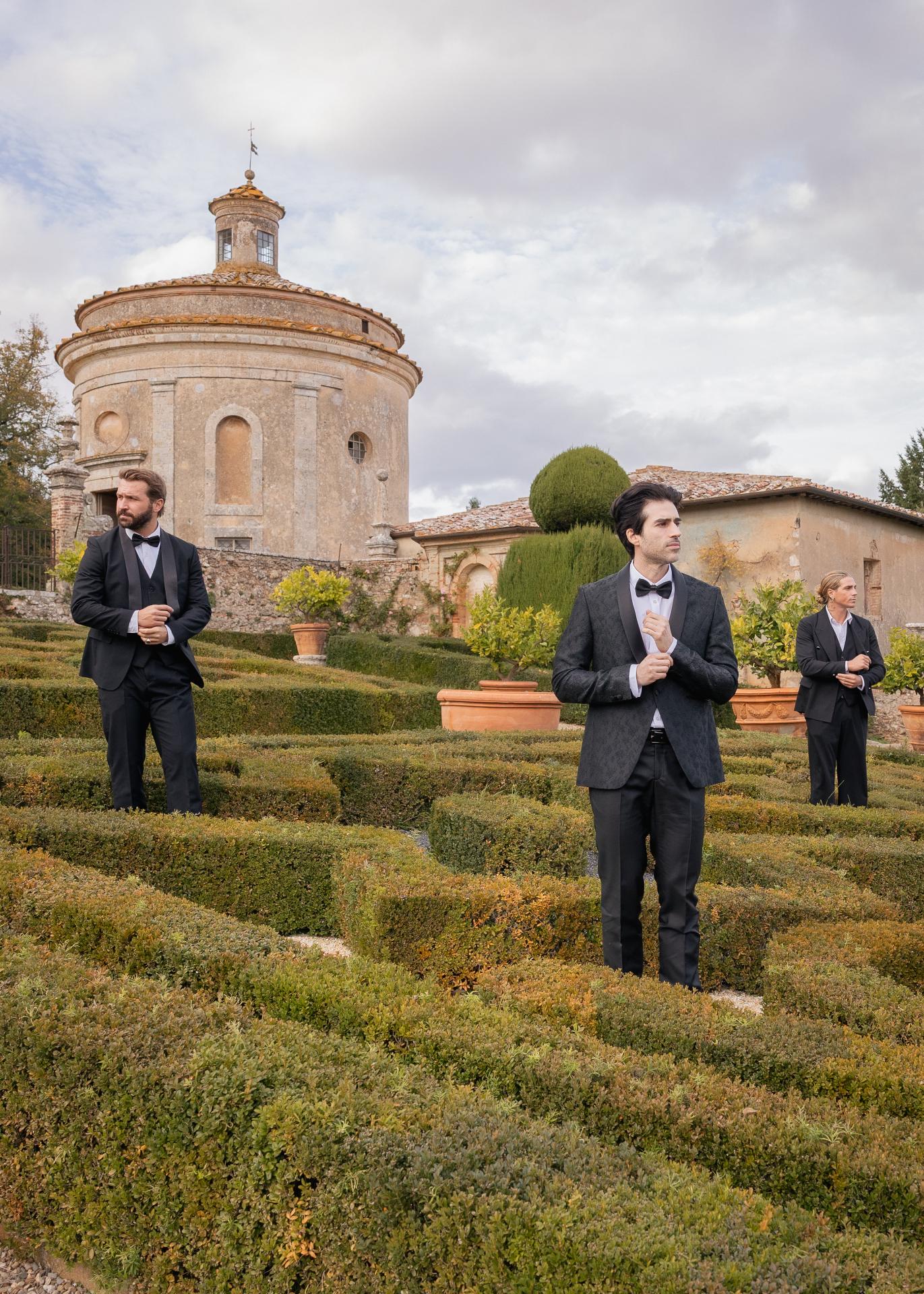 Groom and groomsmen in black tie suits standing in a historic Italian villa garden before a luxury wedding.