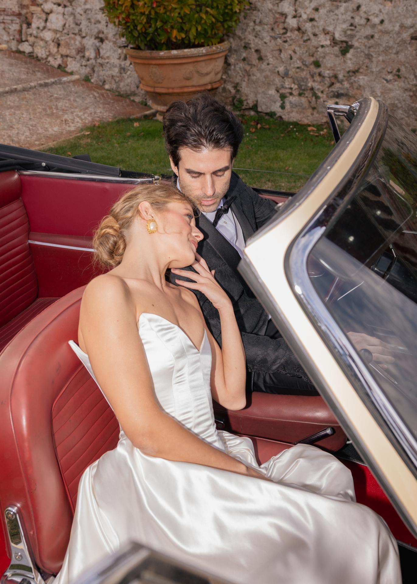 Elegant wedding couple in a vintage convertible car, bride in satin gown and groom in black tuxedo at an Italian villa.