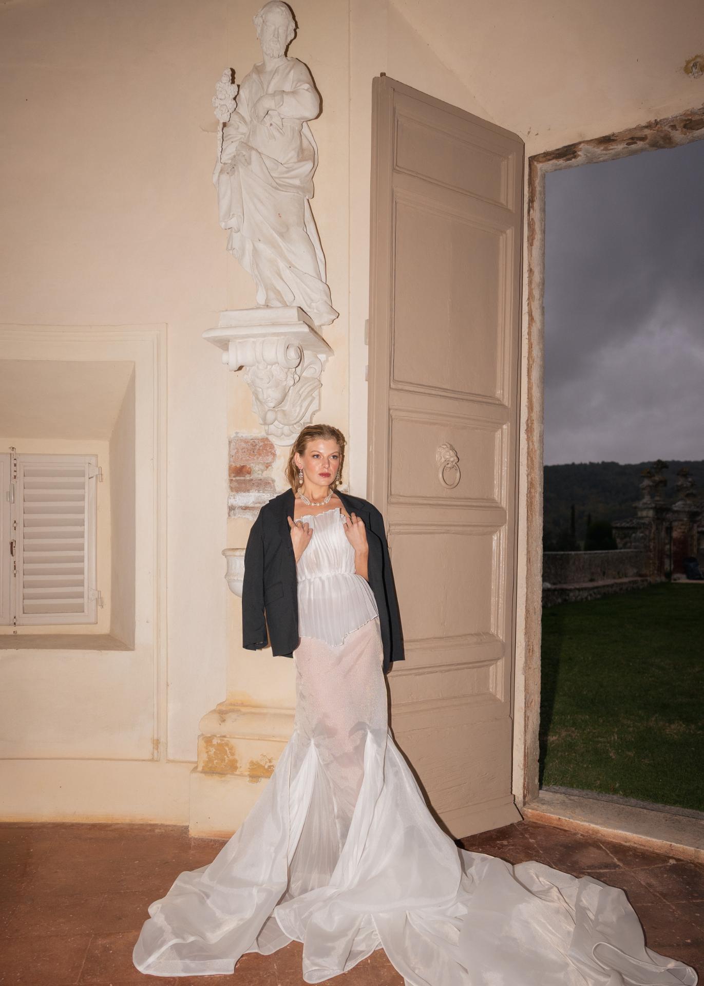 Bride wearing a modern wedding dress and blazer standing at the entrance of an Italian villa with classical sculpture backdrop.