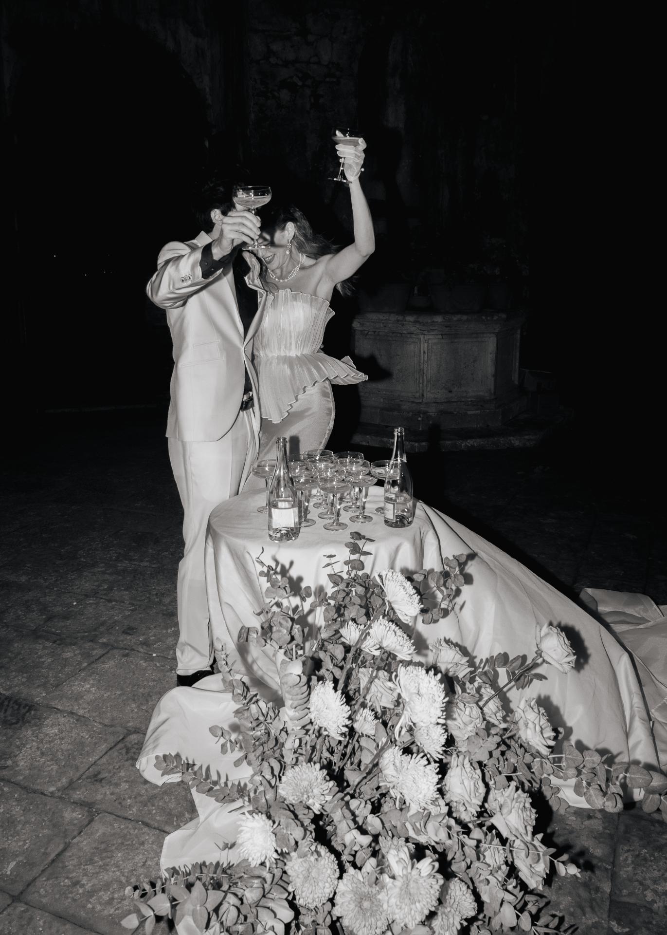 Bride and groom celebrating with champagne at a wedding reception table, captured in a dramatic black and white editorial wedding photograph.