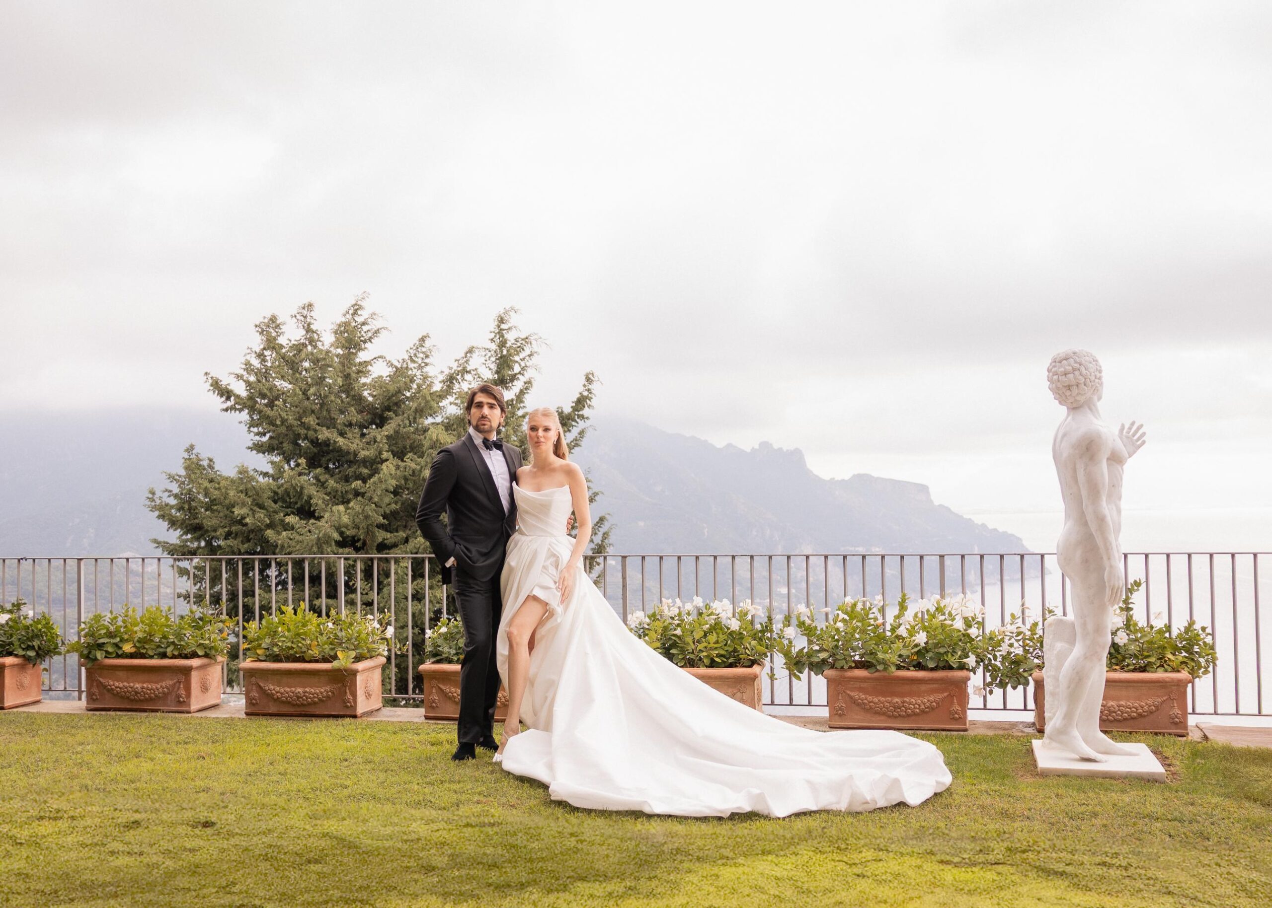 Elegant bride and groom posing on a villa terrace with panoramic Italian mountain views, the bride wearing a modern satin wedding gown with a long train and the groom in a classic black tuxedo.
