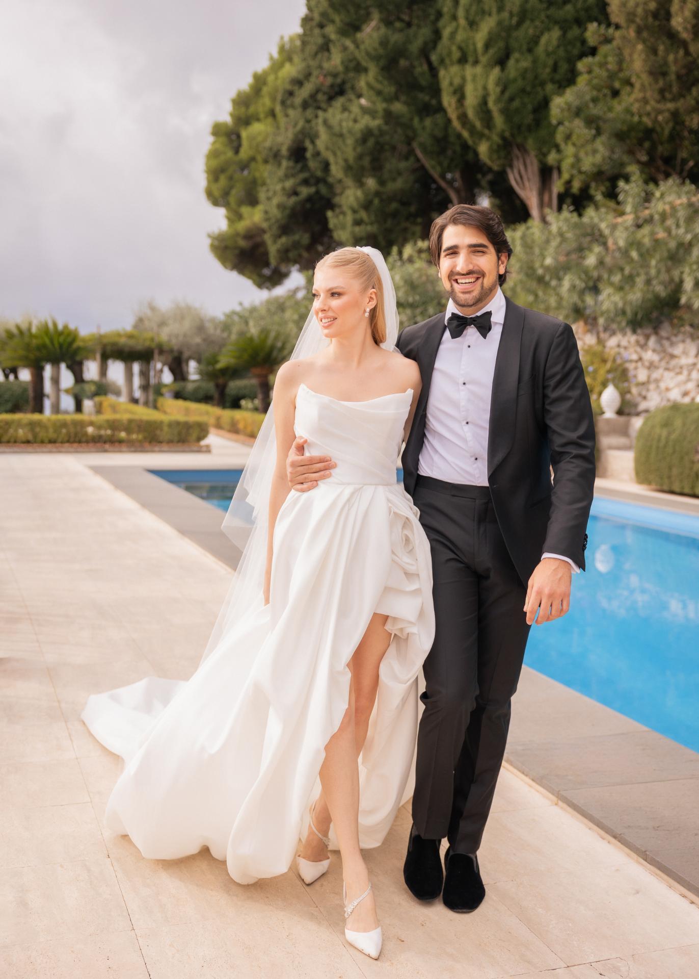 Elegant bride and groom walking hand in hand by a pool at a luxury Italian wedding, wearing a modern bridal gown and classic black tuxedo