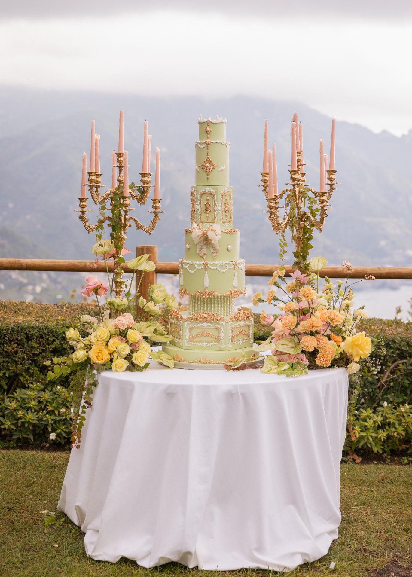 Luxury multi-tier pastel green wedding cake with gold details, florals, and candles, styled outdoors with an Italian mountain view.