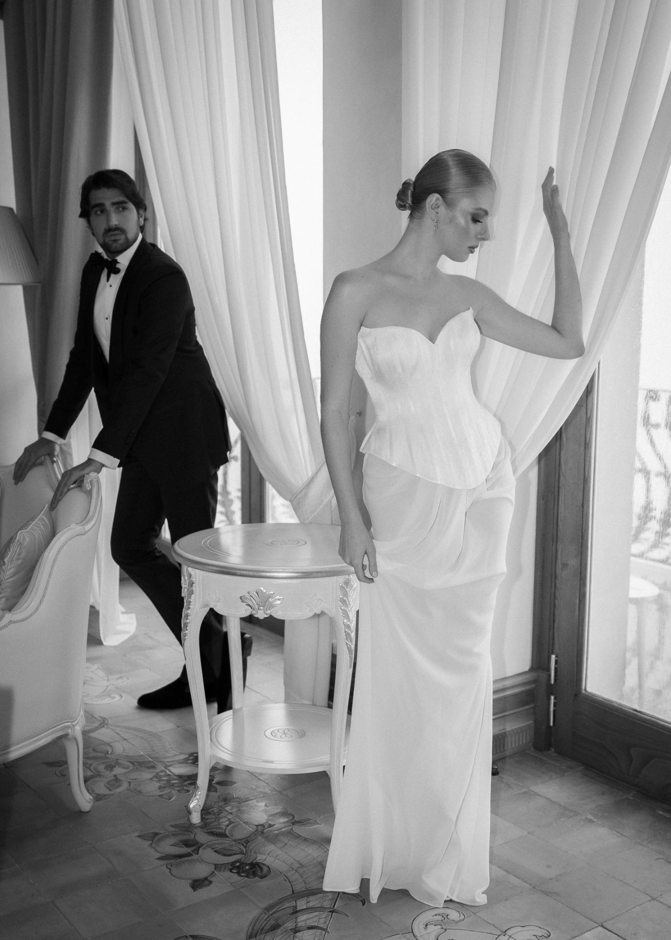 Black and white editorial bridal portrait of a bride in a sculptural corset gown by a window, with groom in tuxedo in the background.