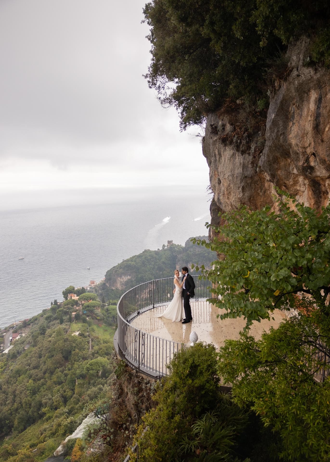Bride and groom standing on a dramatic cliffside balcony overlooking the Amalfi Coast, capturing an intimate destination wedding moment with panoramic sea views and lush Italian landscape.