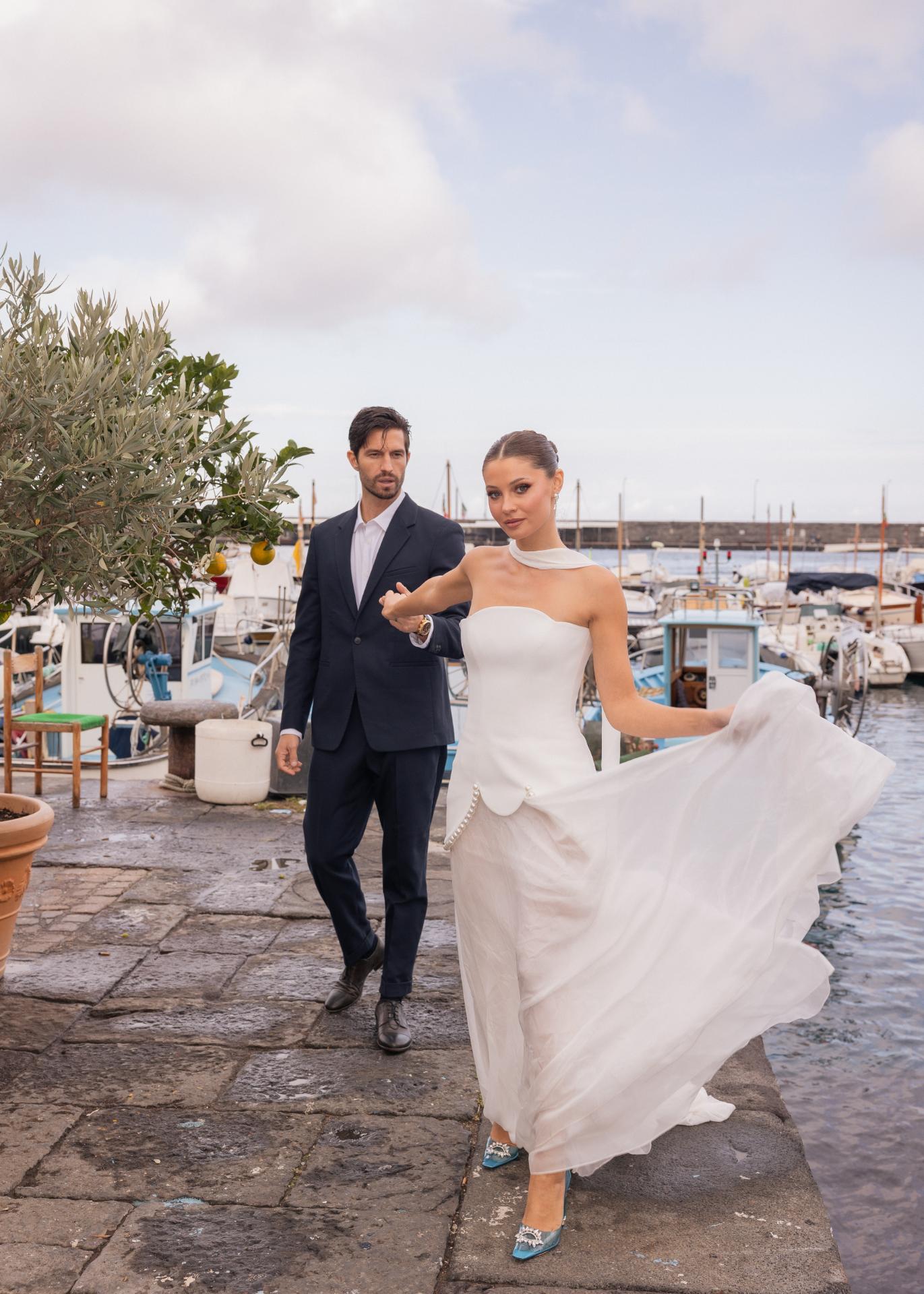 Bride in a modern strapless wedding dress walking along an Italian harbor with the groom, capturing an elegant destination wedding scene.