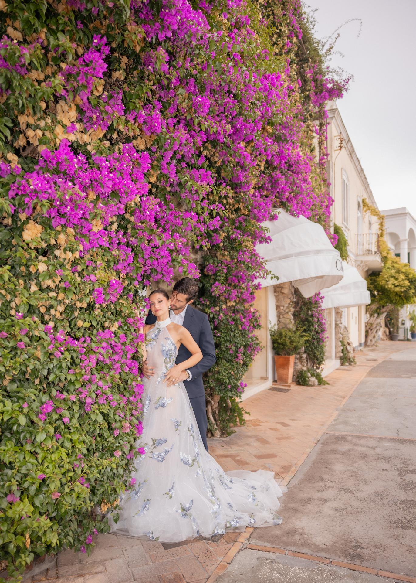 Romantic destination wedding couple embracing beneath vibrant bougainvillea flowers at an elegant Italian villa, the bride wearing a floral embroidered wedding gown and the groom in a classic navy suit.