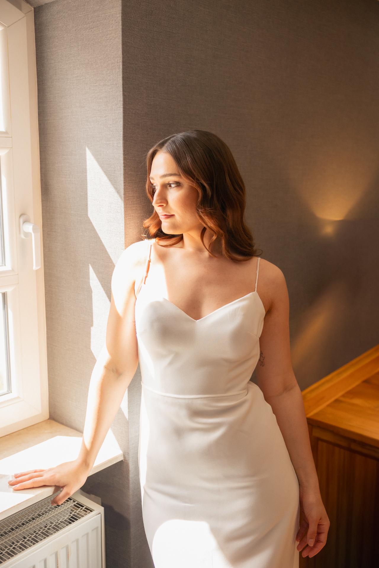 Bride in an elegant wedding dress standing by a window, bathed in warm natural light