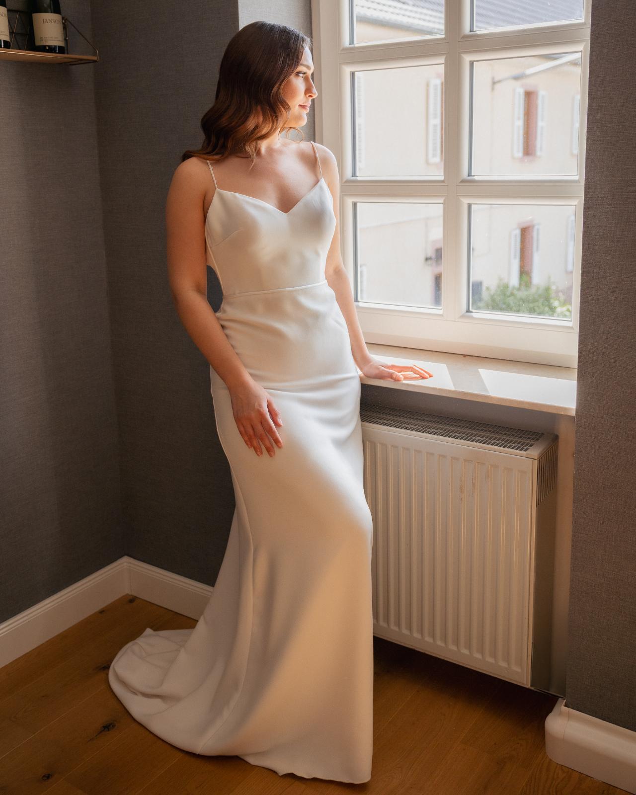 Bride standing by a window in a minimalist wedding dress, softly lit by natural light