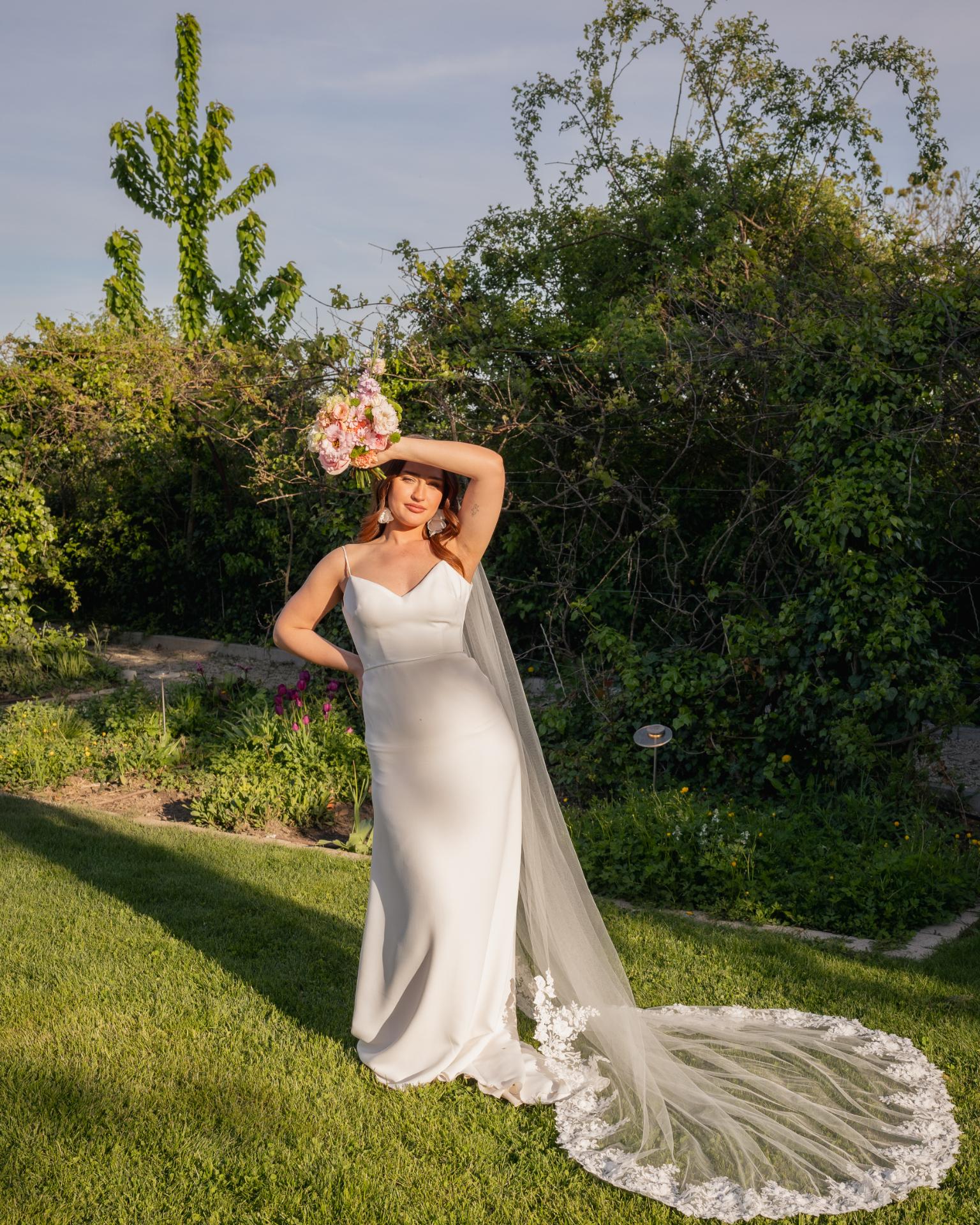 Modern bride in a minimalist wedding dress holding a bouquet, standing in a garden during golden hour with a long flowing veil