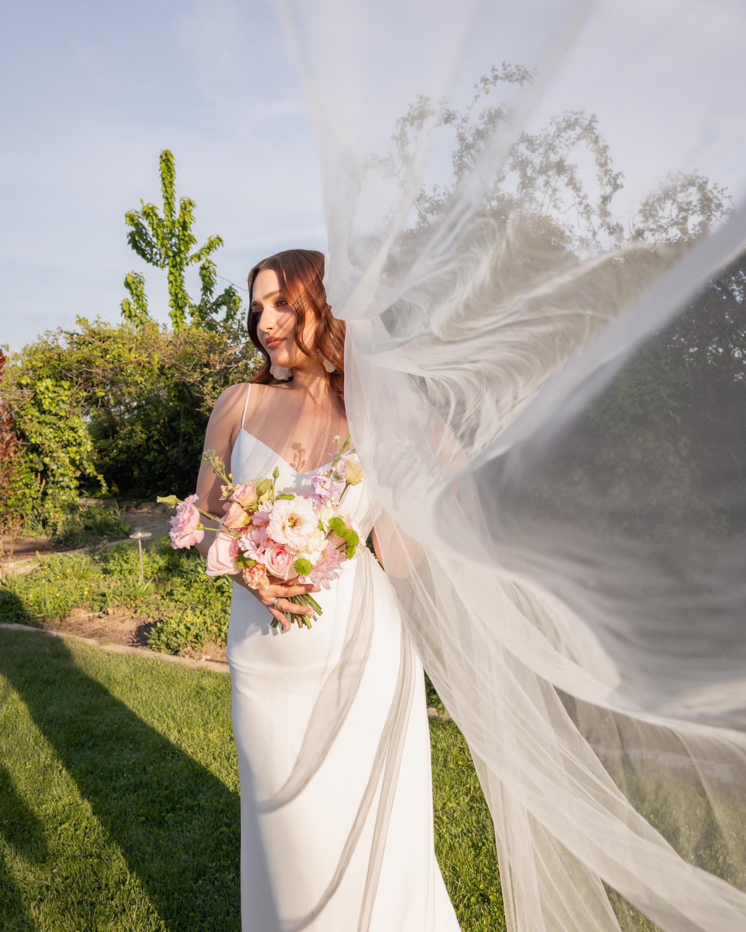 Bride holding a pastel wedding bouquet with a flowing veil in golden hour light, wearing a modern minimalist wedding dress in a garden setting