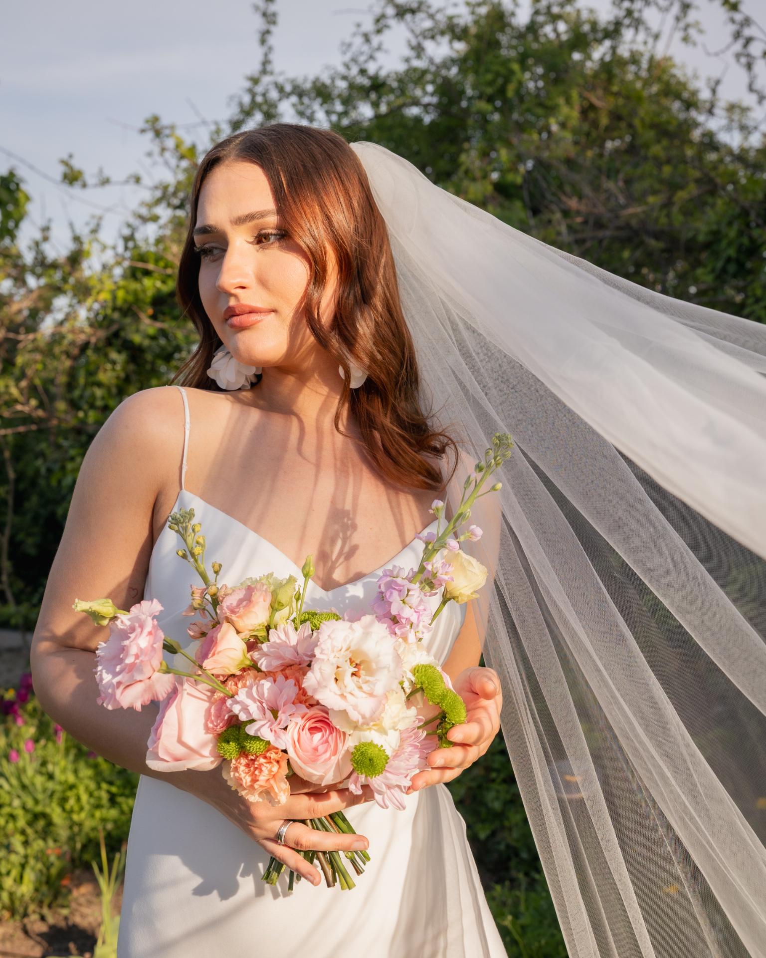 Bride holding a pastel wedding bouquet during golden hour, wearing a flowing veil and a modern minimalist wedding dress in a natural garden setting