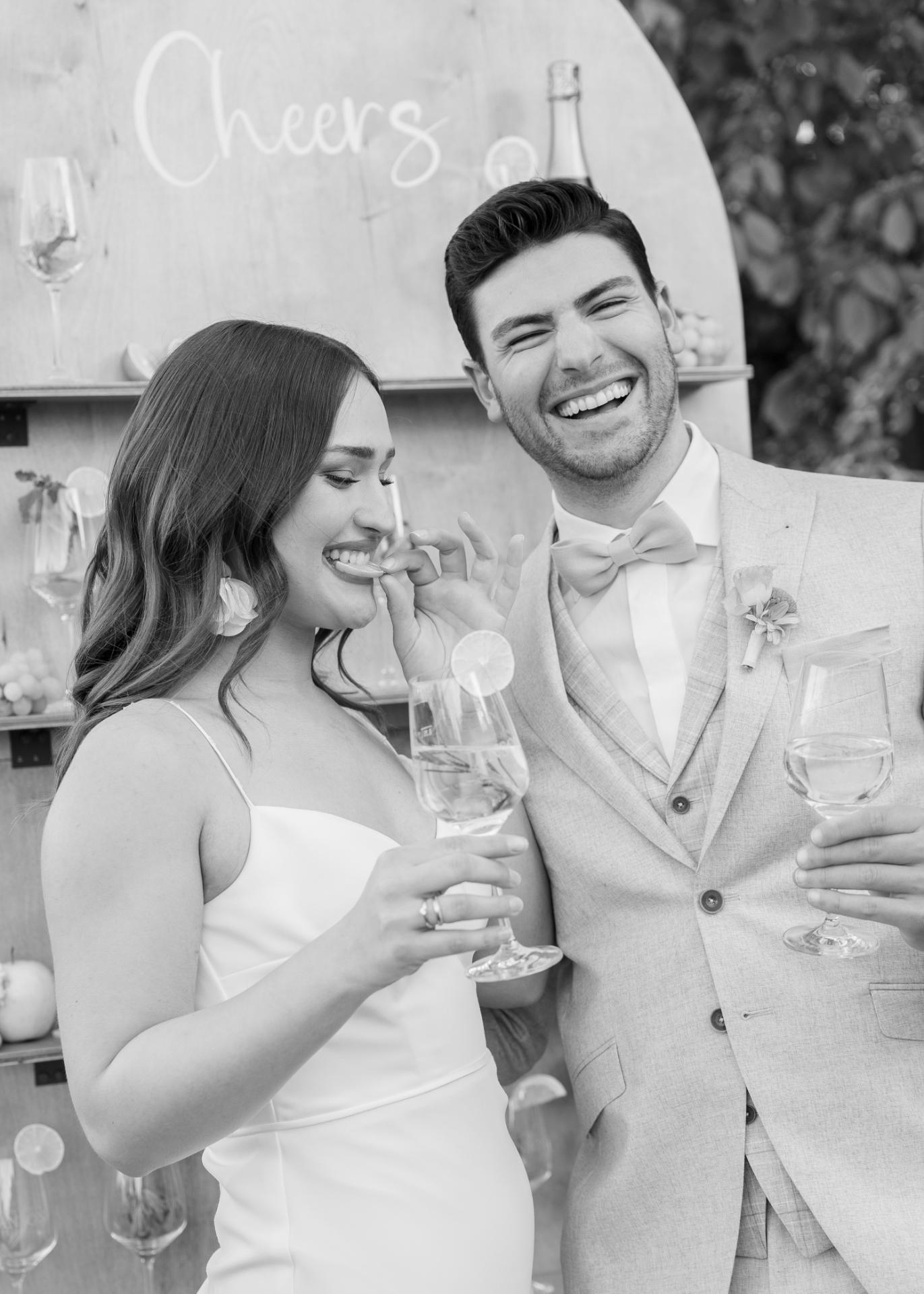 Bride and groom laughing during wedding cocktail hour, holding drinks and sharing an intimate, joyful moment in front of a styled bar backdrop