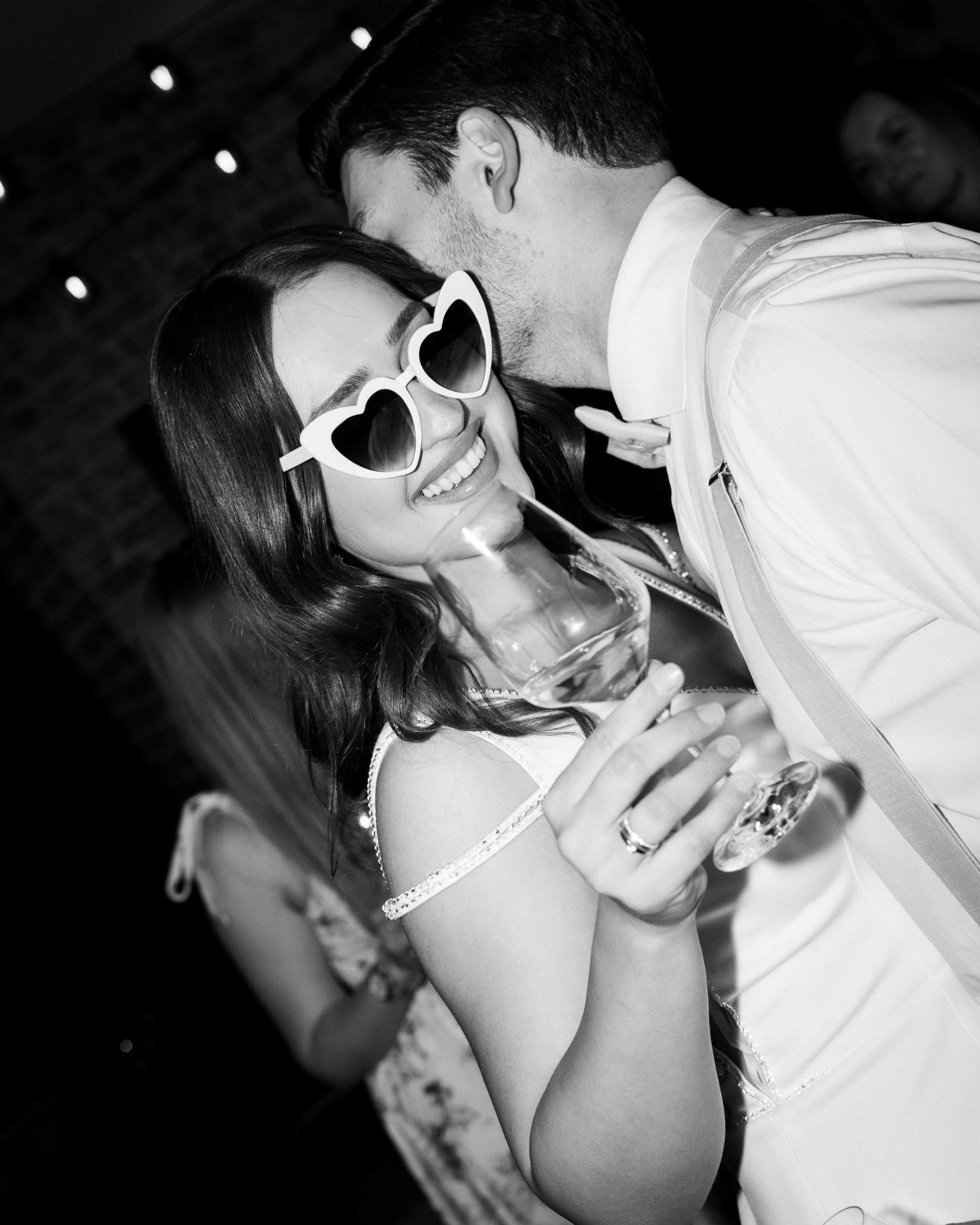 Bride wearing heart-shaped sunglasses during the wedding after party, holding a champagne glass while dancing with the groom