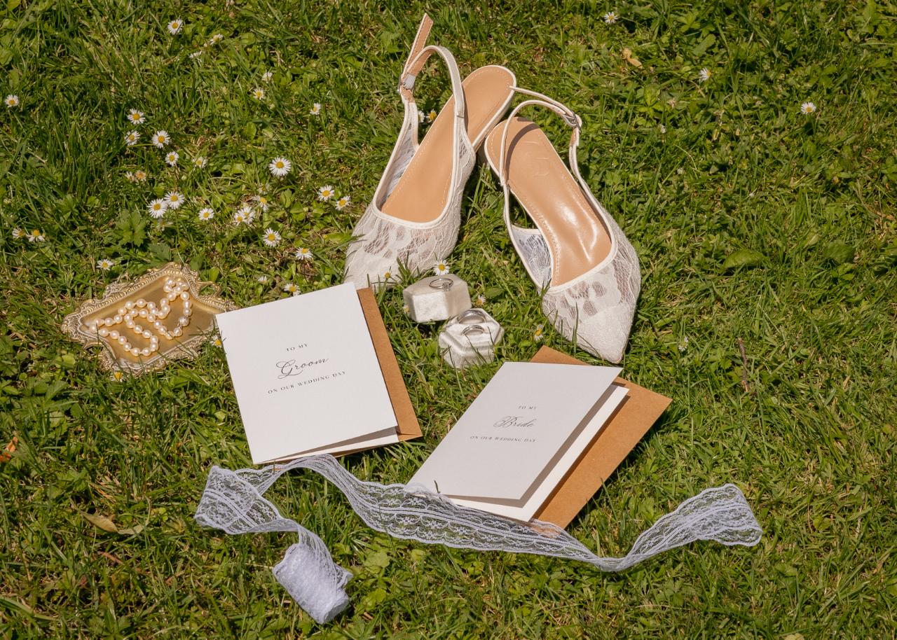 Wedding detail flatlay with bride and groom vow books, lace bridal shoes, wedding rings and pearl accessories arranged on green grass