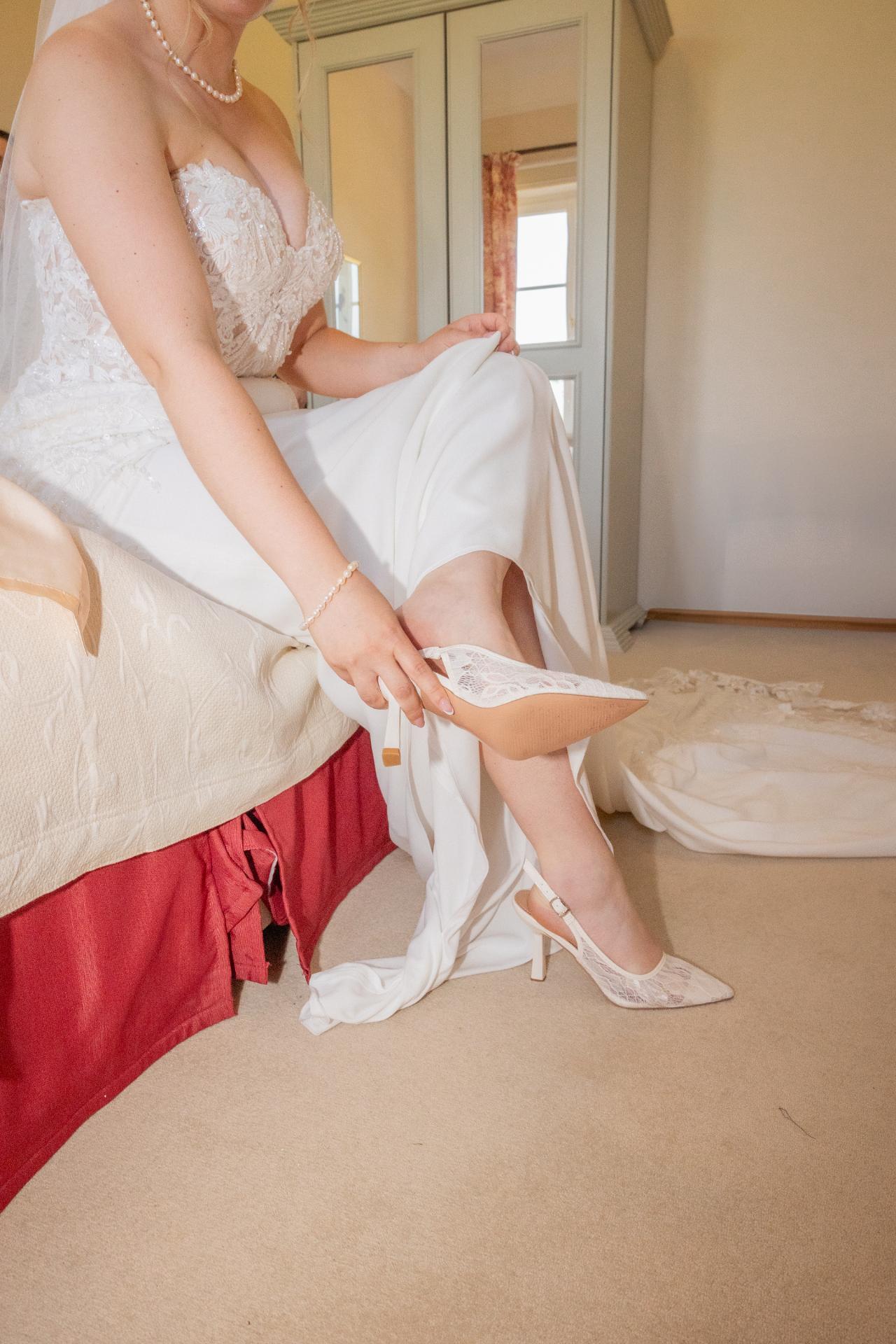 Bride putting on lace wedding shoes during bridal getting ready, wearing an elegant lace wedding dress with pearl jewelry in a softly lit room