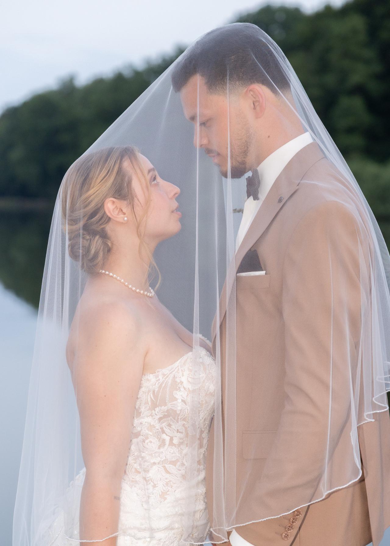 Romantic wedding portrait of bride and groom standing under the veil by a lake, looking at each other