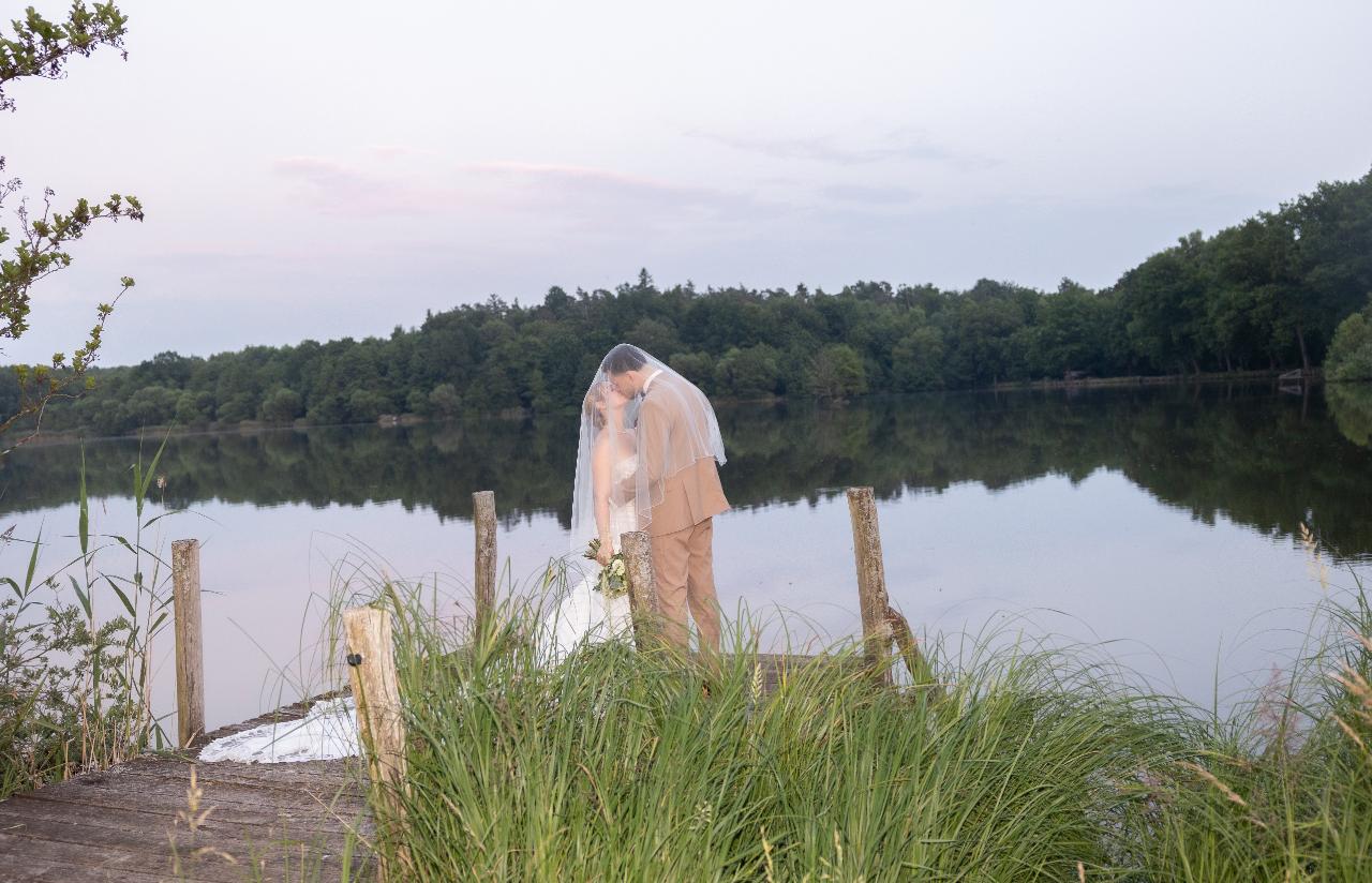 Bride and groom kissing under the veil on a wooden dock by a calm lake at sunset
