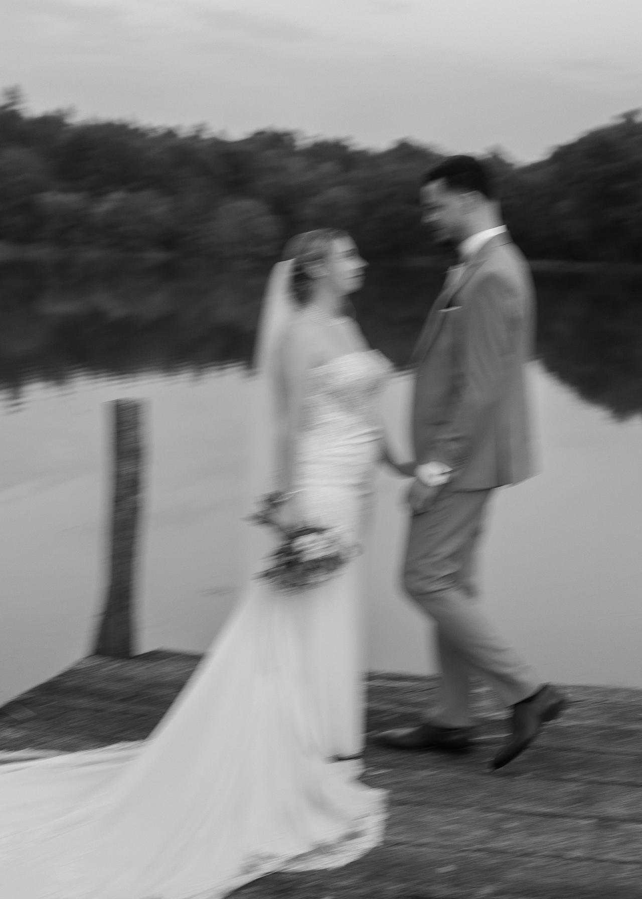 Artistic black and white wedding photo of bride and groom holding hands on a lake dock with motion blur