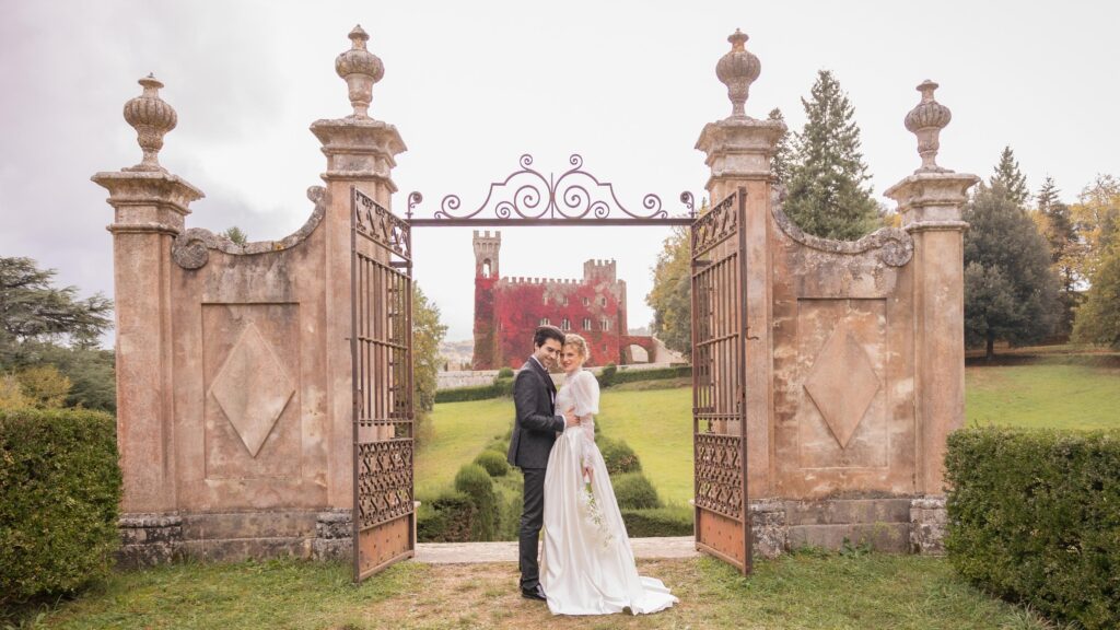Bride and groom standing between the historic gates of Castello di Celsa, Tuscany, with the castle in the background during a romantic destination wedding.