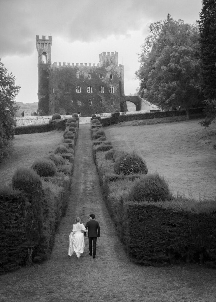 Bride and groom walking hand in hand through the gardens of Castello di Celsa, Tuscany, captured in an elegant black and white wedding photograph.