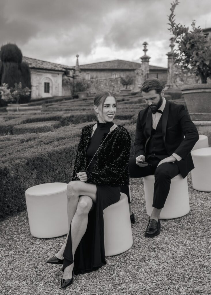 Black and white photo of elegant wedding guests in black tie attire seated in the gardens of Castello di Celsa, Tuscany.