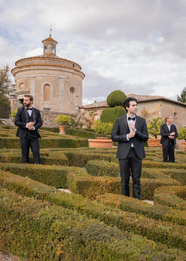 Groom and groomsmen in black tie suits standing in a historic Italian villa garden before a luxury wedding.