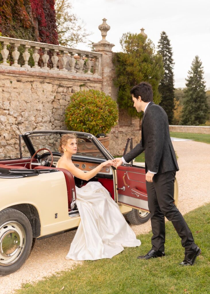 Bride in a satin wedding dress stepping out of a vintage convertible, assisted by the groom at an Italian villa wedding.