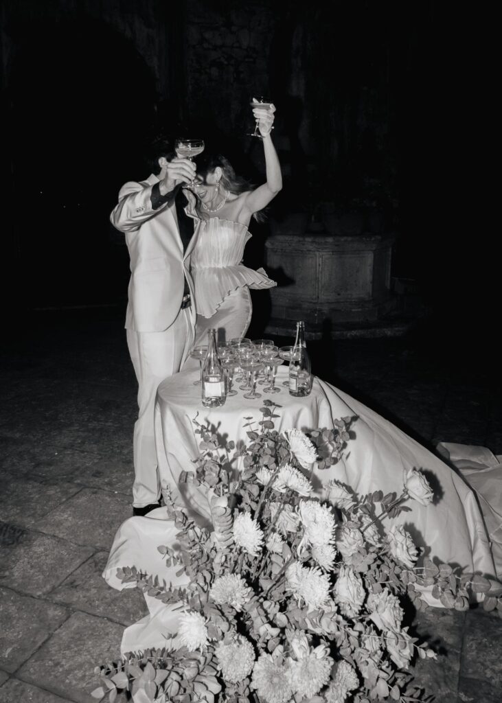 Bride and groom celebrating with champagne at a wedding reception table, captured in a dramatic black and white editorial wedding photograph.