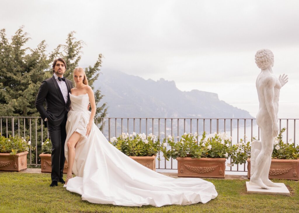 Elegant bride and groom posing on a villa terrace with panoramic Italian mountain views, the bride wearing a modern satin wedding gown with a long train and the groom in a classic black tuxedo.