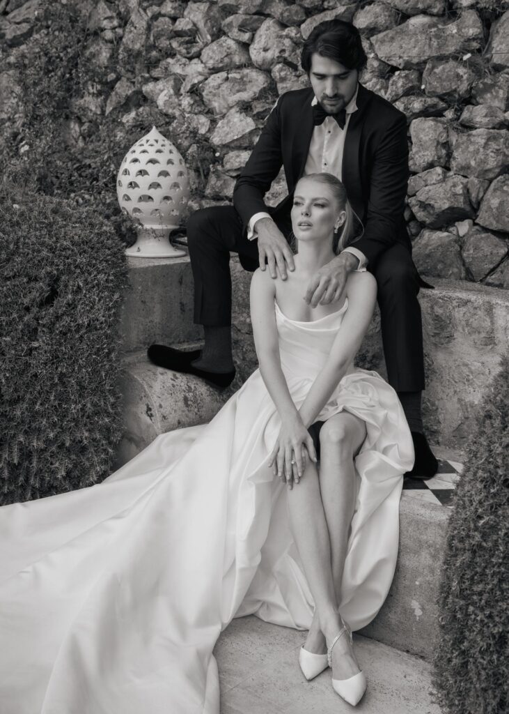 Black and white portrait of an elegant bride seated in a flowing satin wedding gown with the groom in a classic tuxedo behind her at a luxury Italian wedding.
