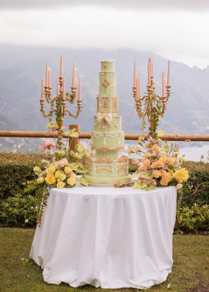 Luxury multi-tier pastel green wedding cake with gold details, florals, and candles, styled outdoors with an Italian mountain view.