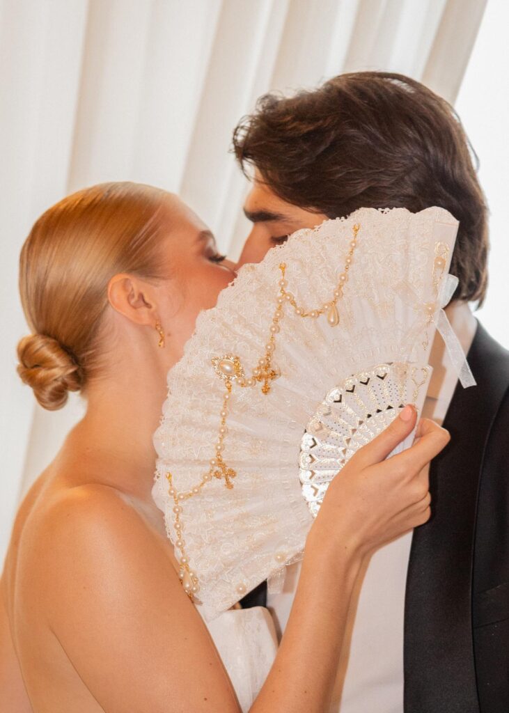 Romantic wedding couple sharing a kiss behind a lace and pearl bridal fan, captured in an elegant editorial wedding portrait.