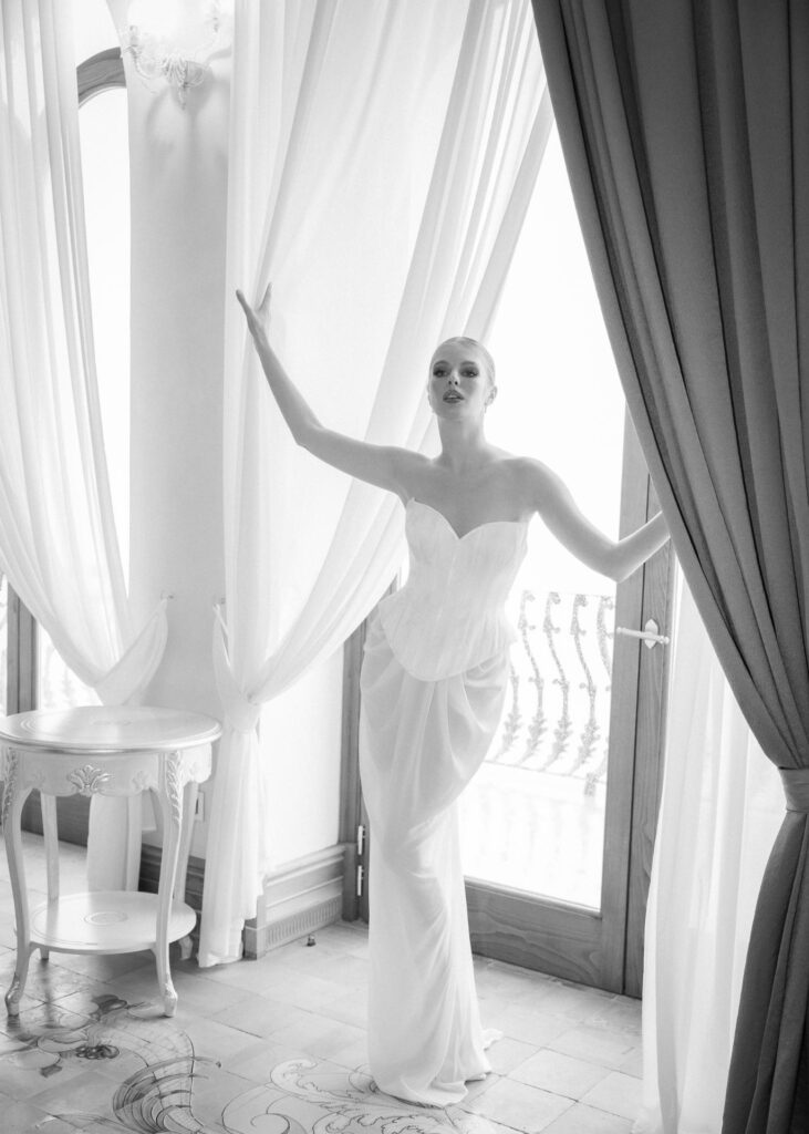 Black and white editorial bridal portrait of a bride wearing a sculptural corset wedding dress, standing between flowing curtains in a luxurious European villa.