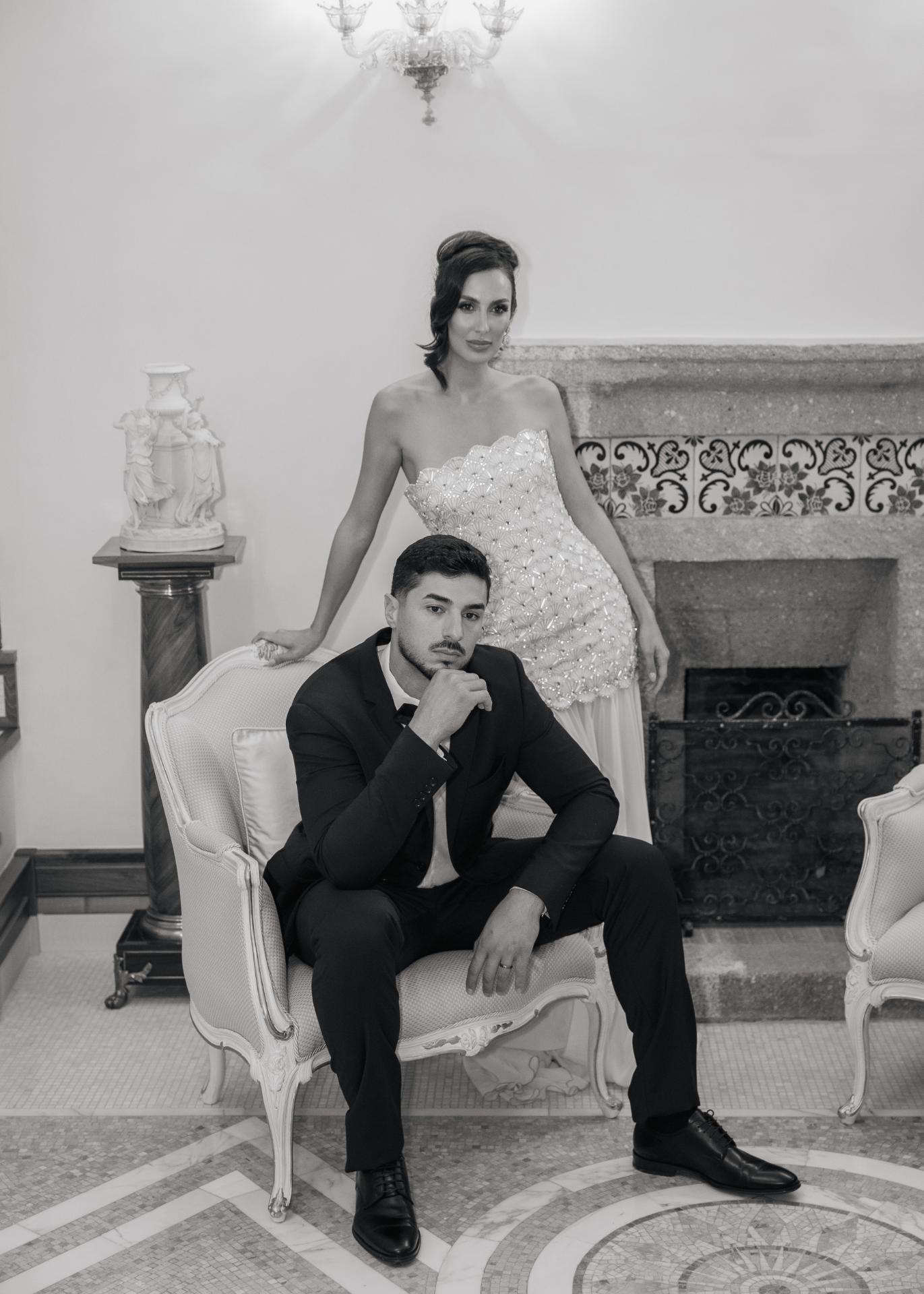 Black and white luxury editorial wedding portrait of an elegant bride in a beaded strapless gown standing behind the groom seated in a classic armchair, styled in a refined European villa interior with timeless, sophisticated wedding aesthetics.