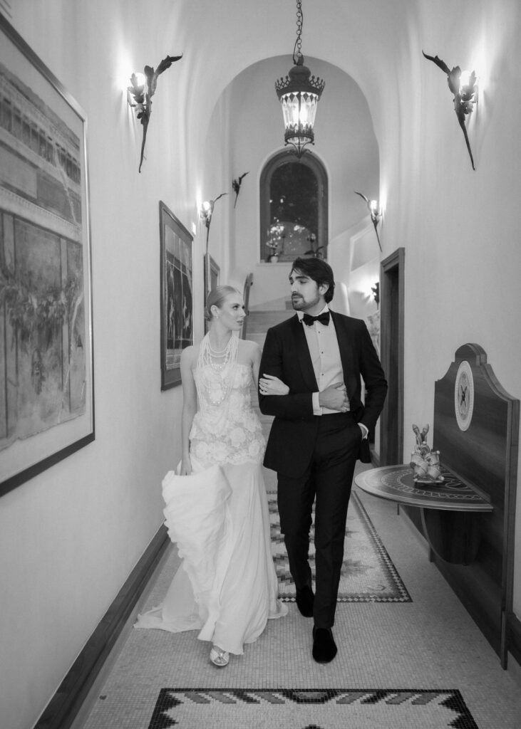 Black and white editorial wedding photograph of an elegant bride and groom walking arm in arm through a grand hallway, featuring a floral lace bridal gown with layered pearls and a classic black-tie tuxedo, captured in a timeless luxury wedding style.