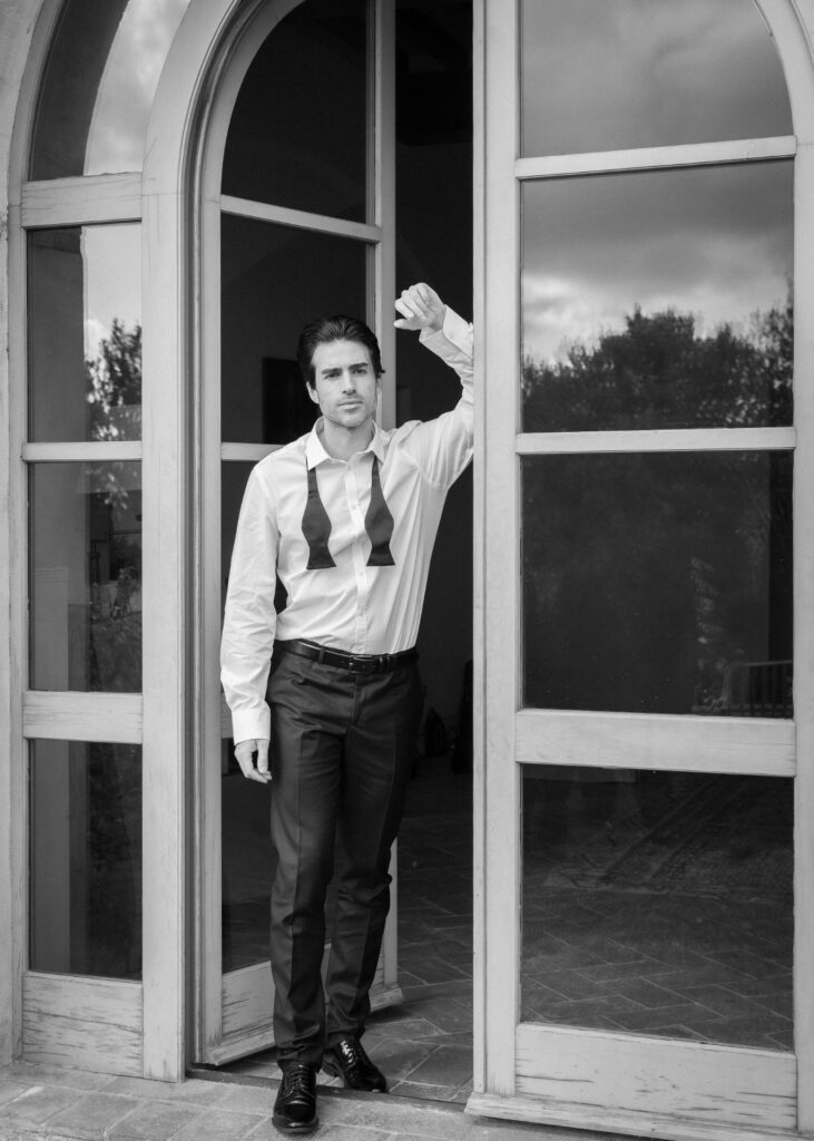 Groom in a black tie wedding look with bow tie undone, standing in an arched window, captured in a timeless black and white editorial wedding portrait.