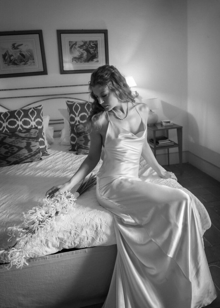 Bride wearing an elegant silk slip wedding dress, seated on a bed with a floral bouquet, captured in a timeless black and white bridal portrait.