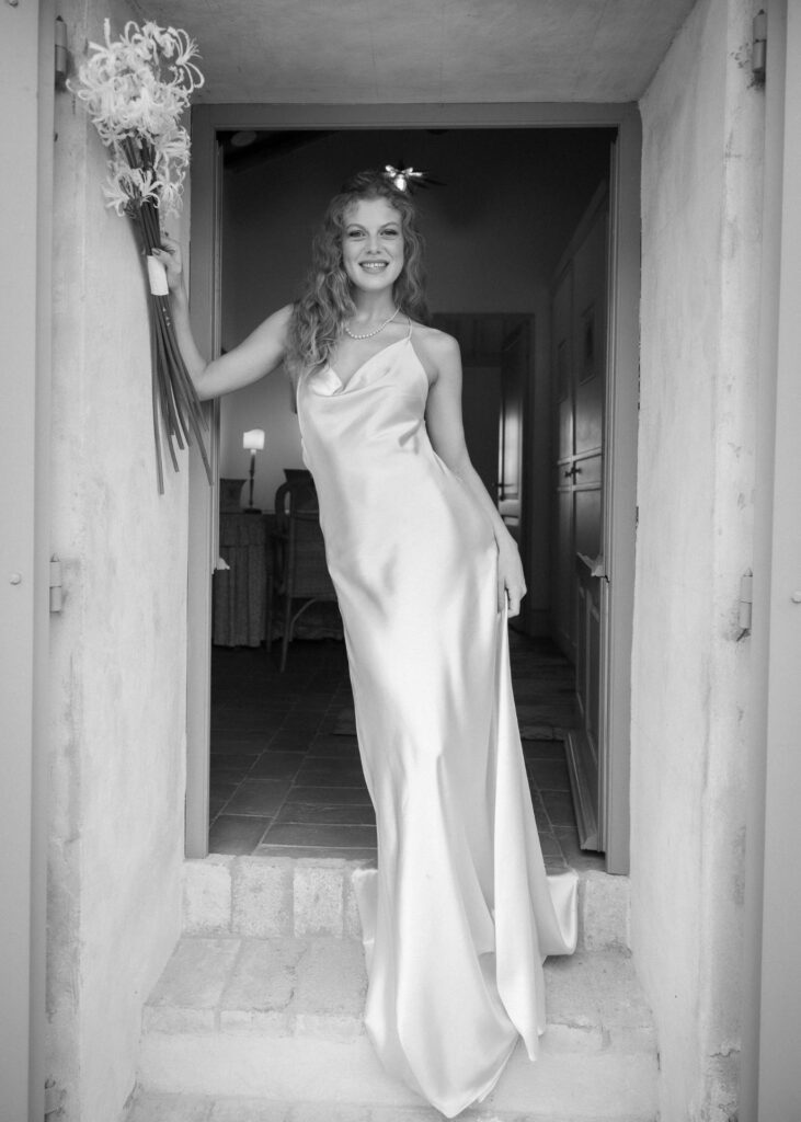 Bride in an elegant silk slip wedding dress standing in a doorway, holding a floral bouquet, captured in a timeless black and white portrait.