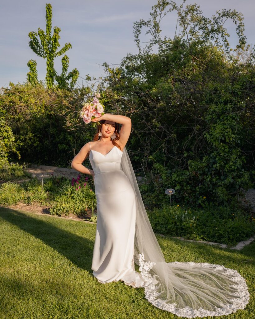 Modern bride in a minimalist wedding dress holding a bouquet, standing in a garden during golden hour with a long flowing veil