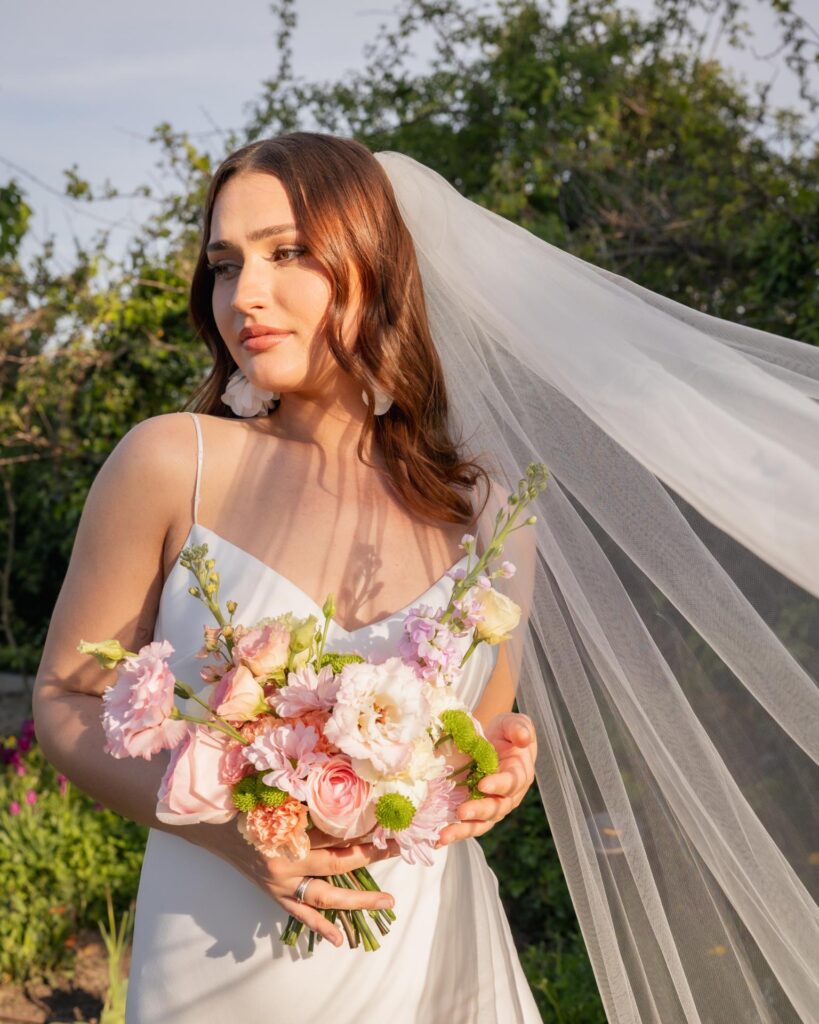 Bride holding a pastel wedding bouquet during golden hour, wearing a flowing veil and a modern minimalist wedding dress in a natural garden setting