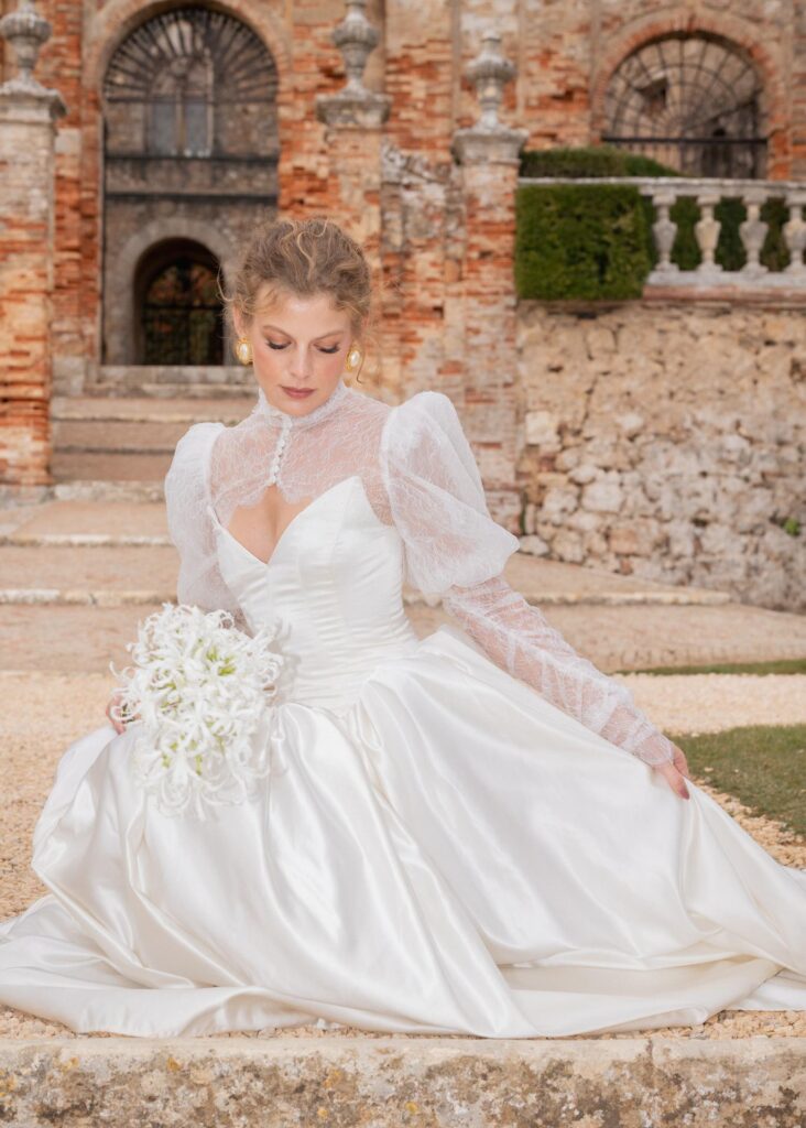 Elegant bridal portrait at Castello di Celsa featuring a bride in a long-sleeve lace wedding dress holding a white bouquet in an Italian castle courtyard